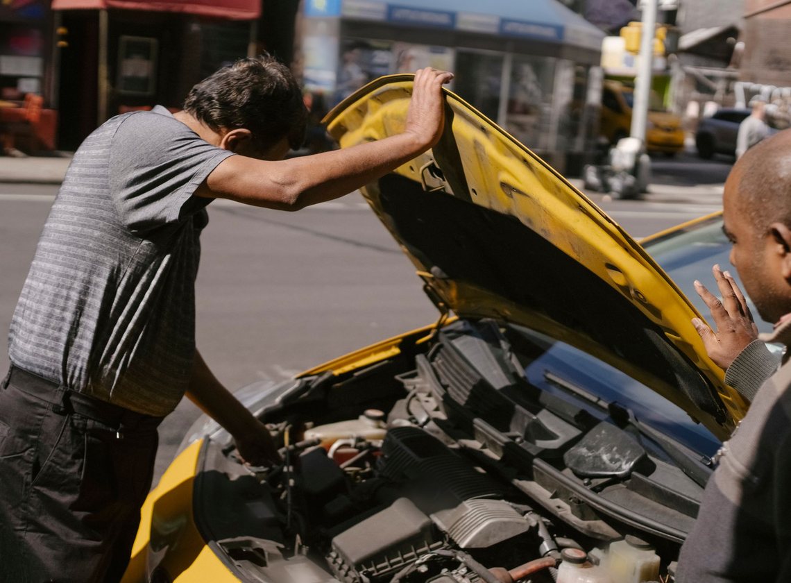 Dois homens conversam diante do motor de um carro amarelo com o capô levantado, numa rua movimentada