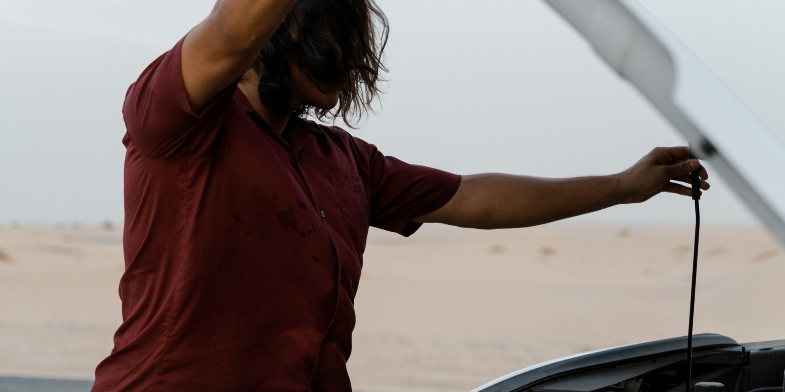 Motorista de camisa vinho puxa a vareta de óleo com o capô aberto numa estrada de areia
